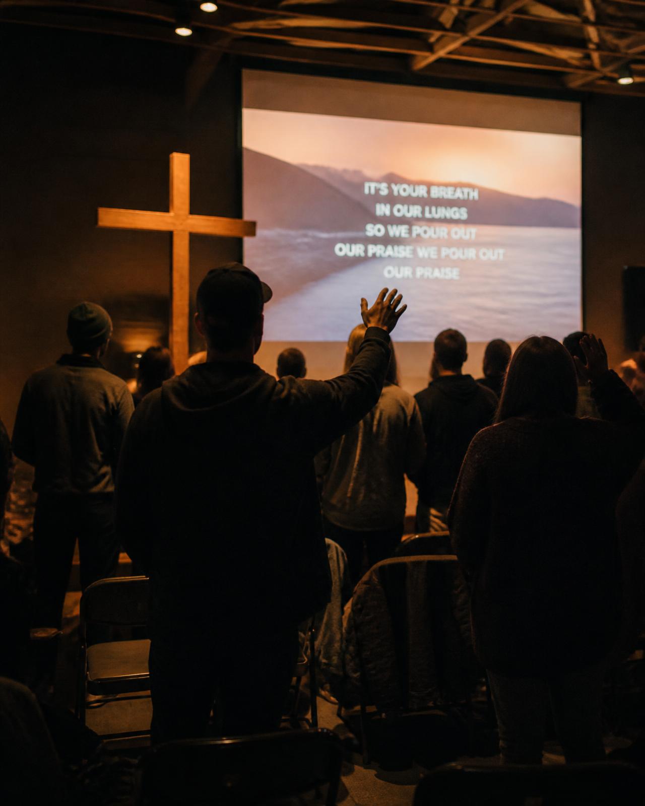 Retreat participants worshipping in a dimly lit room beside a large wooden cross and a worship video on screen